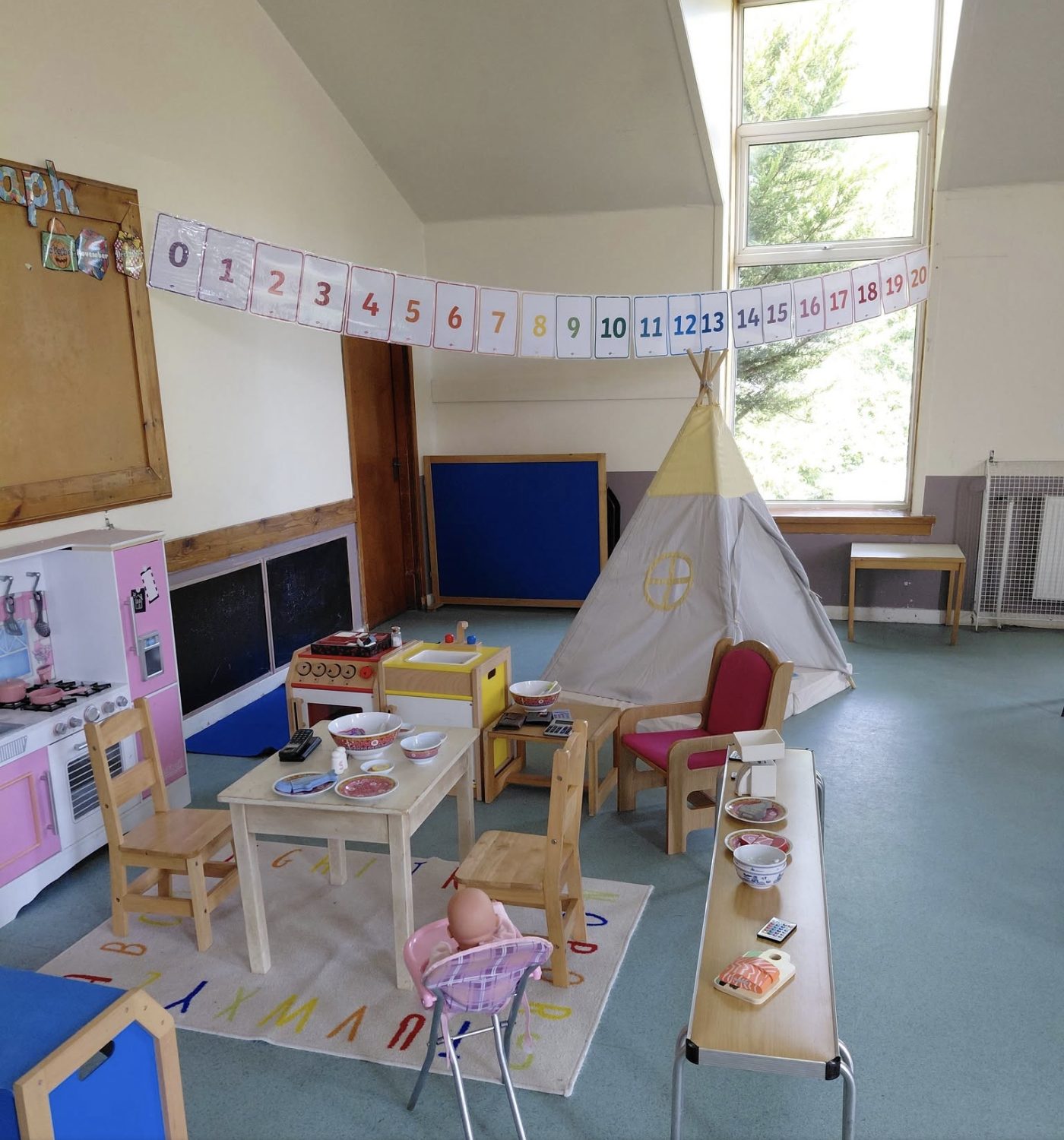 A nursery playroom with a play kitchen, a wooden table and chairs set for pretend lunch, a number line strung overhead, and a teepee reading corner by the window.