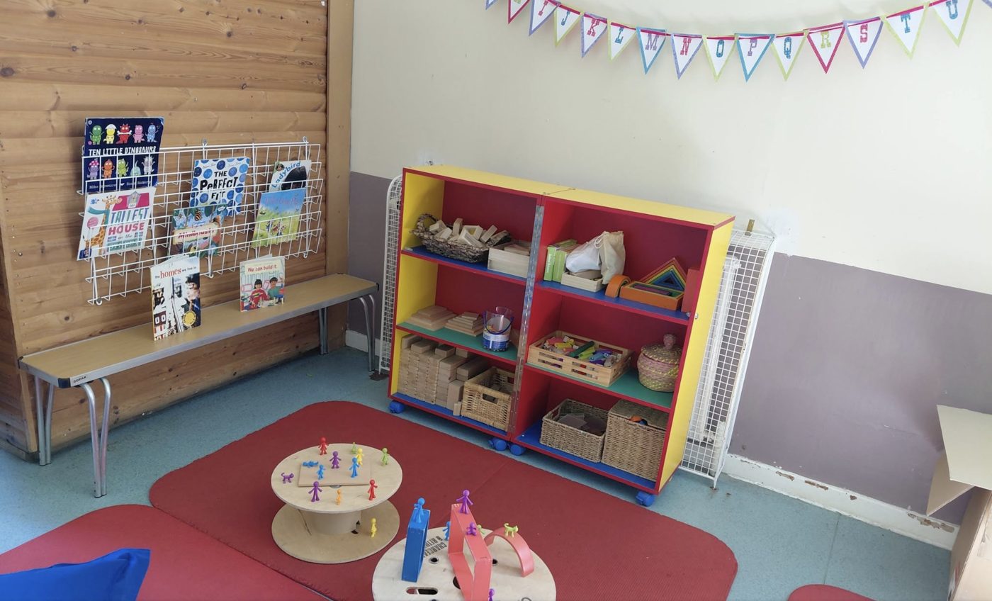 A nursery reading and construction corner with a book rack, storage shelving full of wooden blocks and baskets of toys, and a red play mat with small figures on spinning tables.