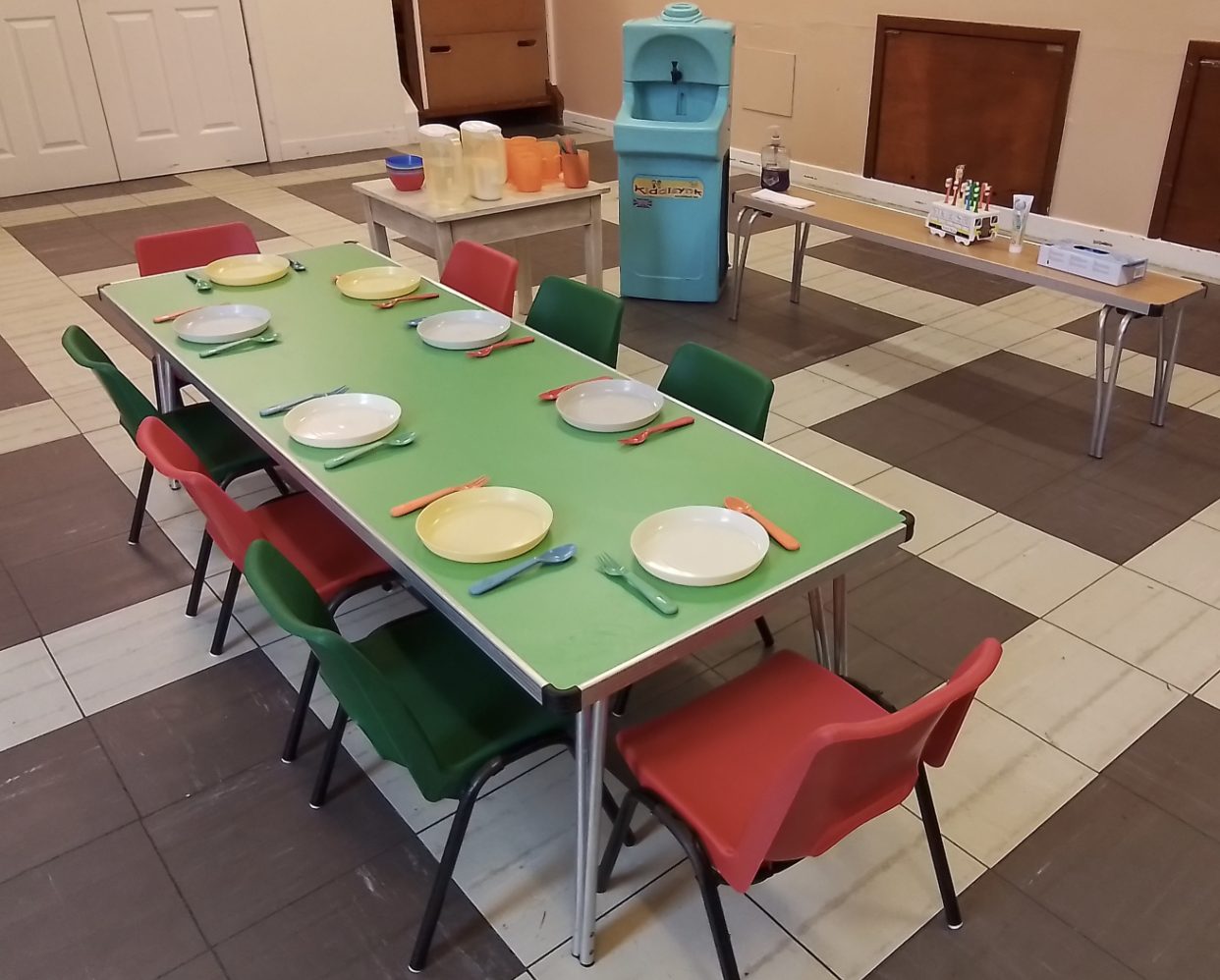 A lunch table set with plates and cutlery for eight children, red and green chairs, and a children's handwashing sink in the background.