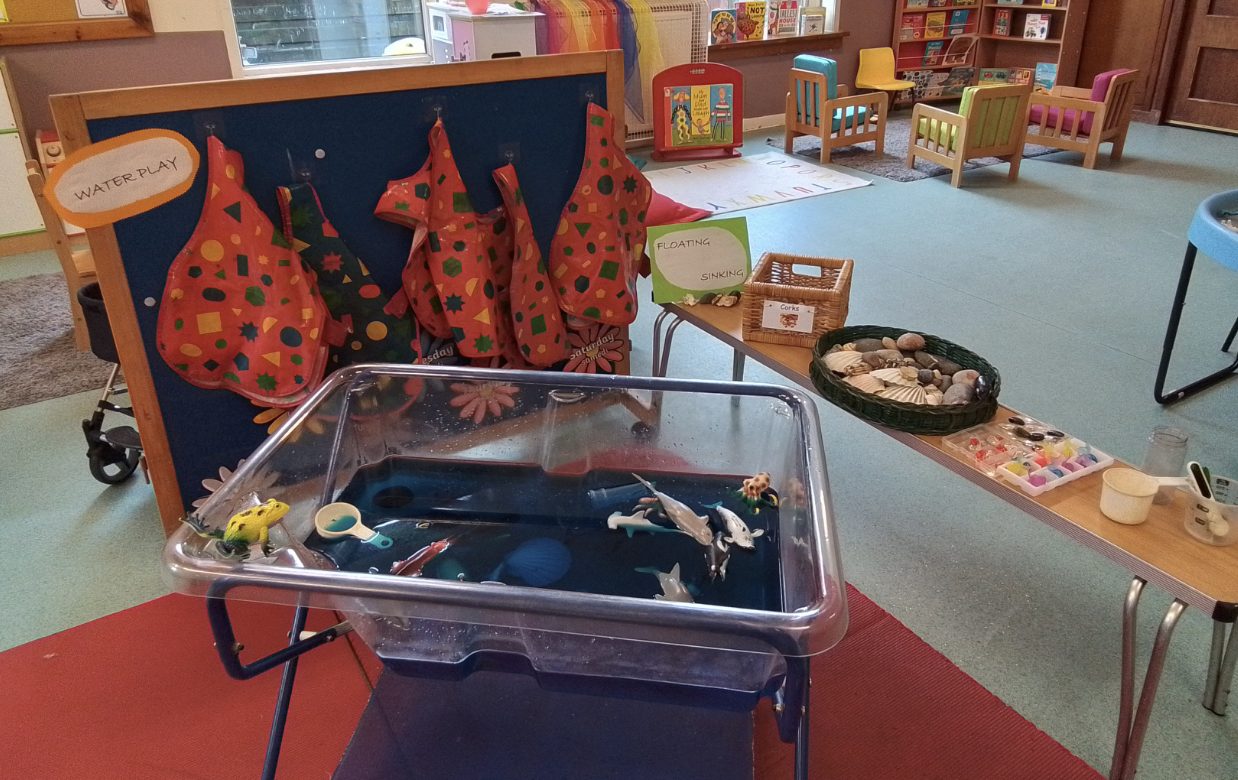 A water play station with a tray of blue water containing toy sea creatures, alongside a floating and sinking activity with shells, stones and corks.